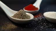 Ceramic spoons filled with different spices and ingredients are arranged on a dark surface. One spoon contains coarse black pepper, another holds bright red paprika, and a third is filled with crystalline white sugar. Small granules are scattered around, enhancing the focus on the textures of the spices.