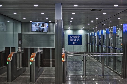 A modern subway station entrance with multiple automated gates featuring green lights. Above the gates, a surveillance camera display screen is mounted, showing live footage. The area is illuminated with ceiling lights, and there's a blue sign indicating platforms 12-13 with directional arrows in both Chinese and English.