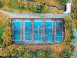 An aerial view of six tennis courts arranged in two rows, surrounded by lush greenery with a path and a small bridge nearby. The courts appear weathered and are mostly blue in color, contrasting with the surrounding autumnal foliage.