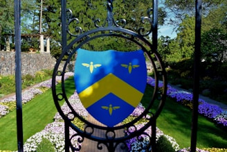 A vibrant garden scene featuring a shield emblem with a yellow chevron and three small yellow symbols against a blue background, mounted on ornamental black ironwork. The garden is lined with neatly arranged pathways, bordered by colorful flowers and lush green grass. In the background, stone architecture and dense trees create a serene outdoor atmosphere.