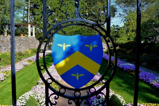 A vibrant garden scene featuring a shield emblem with a yellow chevron and three small yellow symbols against a blue background, mounted on ornamental black ironwork. The garden is lined with neatly arranged pathways, bordered by colorful flowers and lush green grass. In the background, stone architecture and dense trees create a serene outdoor atmosphere.