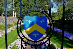 A vibrant garden scene featuring a shield emblem with a yellow chevron and three small yellow symbols against a blue background, mounted on ornamental black ironwork. The garden is lined with neatly arranged pathways, bordered by colorful flowers and lush green grass. In the background, stone architecture and dense trees create a serene outdoor atmosphere.