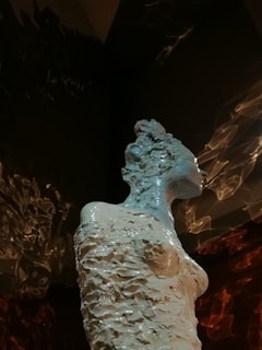 A textured sculpture of a human figure with a rough, fragmented surface, standing against a dark background. The lighting highlights the intricacies of the sculpture, casting dramatic shadows.