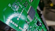 A close-up view of a green printed circuit board with various electronic components, including a black integrated circuit chip in the center. The circuit board has multiple silver solder points and white printed labels such as 'Driver'.
