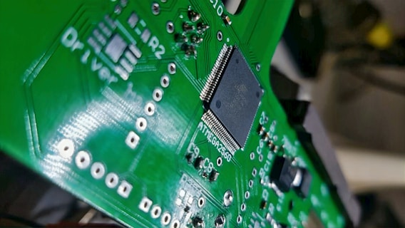 A close-up view of a green printed circuit board with various electronic components, including a black integrated circuit chip in the center. The circuit board has multiple silver solder points and white printed labels such as 'Driver'.