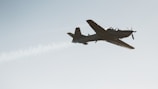 A single-engine aircraft is captured in flight against a clear sky, trailing a visible stream of white smoke. The aircraft appears to be small and possibly designed for aerobatics or training.