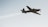 A single-engine aircraft is captured in flight against a clear sky, trailing a visible stream of white smoke. The aircraft appears to be small and possibly designed for aerobatics or training.