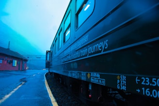 A train is parked alongside a platform in a picturesque, mountainous area. The side of the train features promotional text about scenic train journeys. The sky is overcast, casting a cool blue tone across the scene. A small red building is visible on the left side of the image, with tracks leading into the distance.