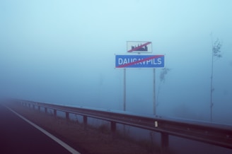 A foggy scene with a road sign indicating the end of a location. The road sign has the name 'DAUGAVPILS' crossed out with a red line. The atmosphere is misty, with limited visibility beyond the sign. To the left is a metal guardrail bordering the edge of a road.