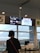 A person is standing in front of a departure gate at an airport, looking at the flight information display screen. The screen shows details for a flight to Taipei, operated by China Airlines and Japan Airlines. The environment is inside an airport terminal, with large windows and some visible seating.
