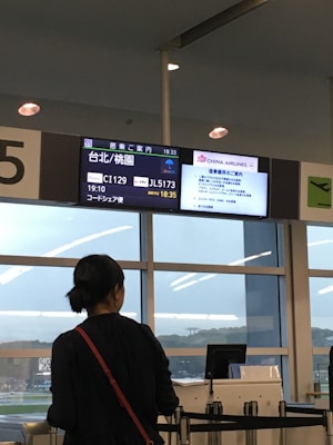 A person is standing in front of a departure gate at an airport, looking at the flight information display screen. The screen shows details for a flight to Taipei, operated by China Airlines and Japan Airlines. The environment is inside an airport terminal, with large windows and some visible seating.