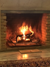 A cozy fireplace with a warm fire burning. The flames rise around logs placed on a metal grate within a brick fireplace. The light from the fire reflects on the floor, creating a warm and inviting atmosphere.
