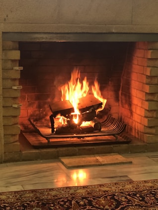 A cozy fireplace with a warm fire burning. The flames rise around logs placed on a metal grate within a brick fireplace. The light from the fire reflects on the floor, creating a warm and inviting atmosphere.