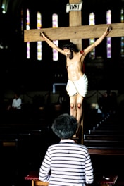 A person with curly hair, wearing a striped shirt, is kneeling in front of a crucifix with a figure of Jesus. The setting appears to be a dimly lit church with stained glass windows visible in the background.