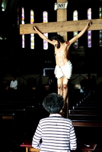 A person with curly hair, wearing a striped shirt, is kneeling in front of a crucifix with a figure of Jesus. The setting appears to be a dimly lit church with stained glass windows visible in the background.