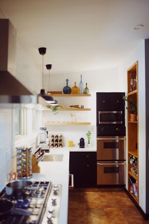 A modern, bright kitchen features stainless steel appliances, including a double oven and a stove with pots and pans. White countertops and tiled walls create a clean look. Open shelving holds decorative vases and plants, while a wine rack and coffee machine are placed next to the sink. Warm lighting illuminates the room, enhancing the cozy atmosphere.