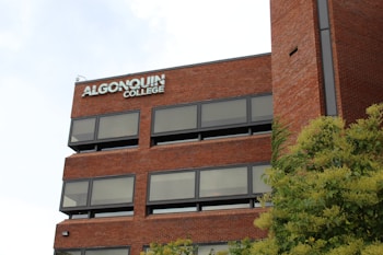 A multi-story brick building with numerous rectangular windows and signage displaying 'Algonquin College' on the upper part. There is some greenery, including trees, visible in the foreground.