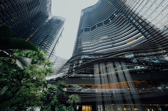 High-rise buildings with a modern architectural design featuring curved lines and a glass facade. Lush greenery is visible in the foreground, creating a contrast with the urban landscape. A water feature cascades down from the structure, adding a dynamic element.