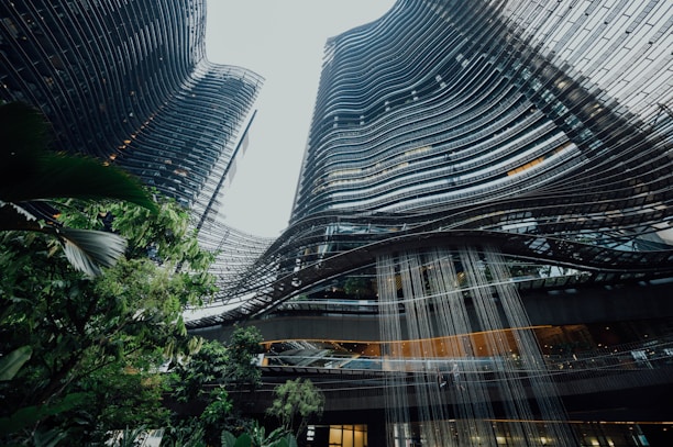 High-rise buildings with a modern architectural design featuring curved lines and a glass facade. Lush greenery is visible in the foreground, creating a contrast with the urban landscape. A water feature cascades down from the structure, adding a dynamic element.
