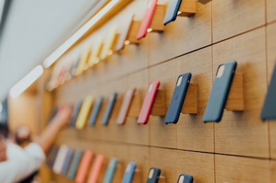 A wall display showcasing an array of colorful smartphone cases mounted on a wooden panel, arranged in a neat and orderly manner. The cases vary in colors including blue, red, and yellow. A person's hand is reaching towards one of the cases.