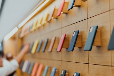 A wall display showcasing an array of colorful smartphone cases mounted on a wooden panel, arranged in a neat and orderly manner. The cases vary in colors including blue, red, and yellow. A person's hand is reaching towards one of the cases.