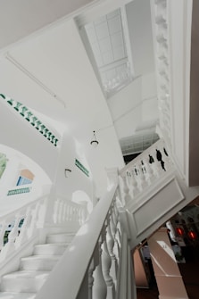 A winding white staircase featuring intricately designed banisters, situated in a bright architectural space with green ornamental details. The ceiling is adorned with decorative tile patterns, and there are small hanging lights. The building has an open and airy feel, with sunlight casting shadows across the surfaces.
