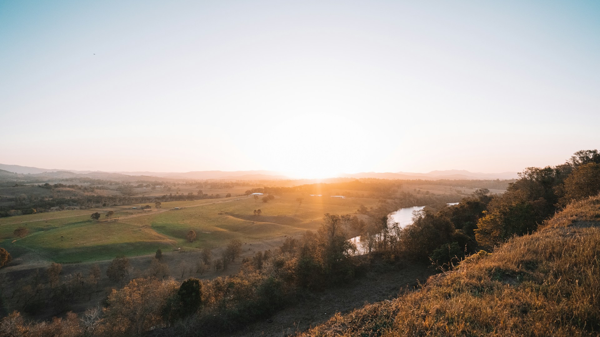 A serene landscape with a soft sunrise illuminating gentle hills and a calm river.