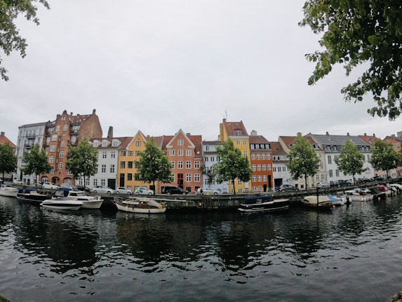 A picturesque canal lined with colorful historic buildings, featuring architectural styles typical of Northern Europe. Several small boats are moored along the canal, reflecting softly in the water. Trees are interspersed between the buildings, adding greenery to the scene. The sky is overcast, suggesting a calm and cool day.