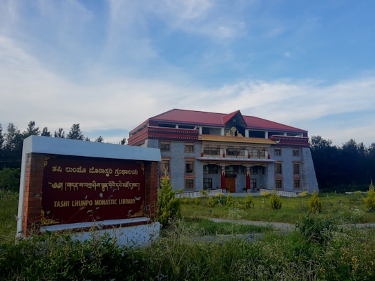 A large monastic library building with a red roof and intricate golden decorations stands prominently in a grassy area. In the foreground, there is a sign indicating the name of the library, which is surrounded by plants. The sky is blue with some clouds and trees line the background.