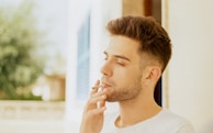 A man with short hair and a beard is standing outdoors, smoking a cigarette. He appears relaxed, with his eyes closed, surrounded by a soft-focus background of greenery and architectural elements.