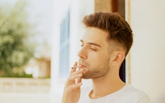 A man with short hair and a beard is standing outdoors, smoking a cigarette. He appears relaxed, with his eyes closed, surrounded by a soft-focus background of greenery and architectural elements.