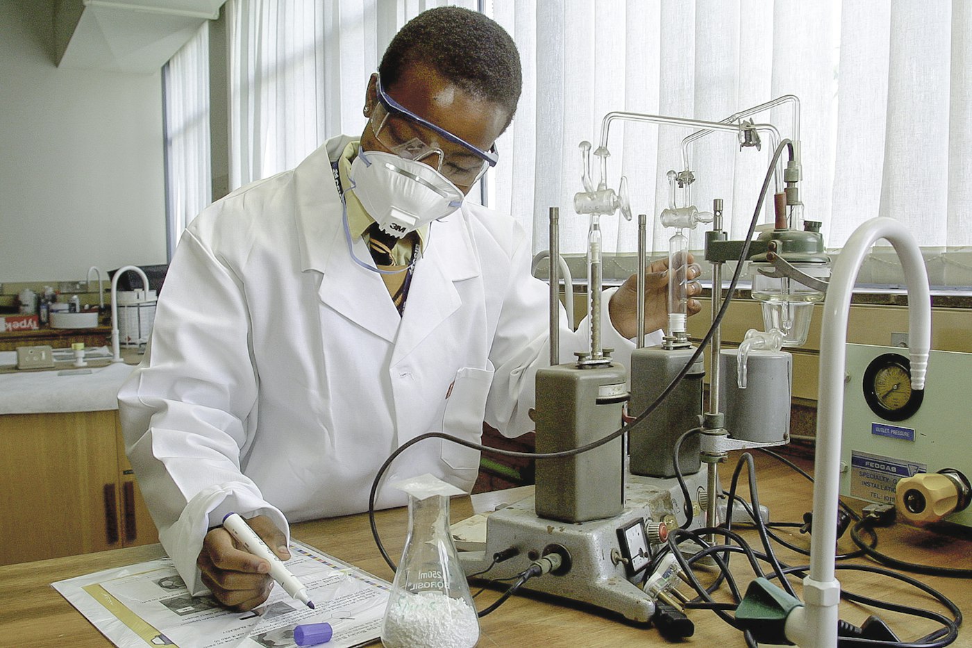 A young researcher in safety glasses and a mask taking notes at a laboratory bench