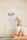A minimalistic calendar page displaying September is clipped to the wall with wooden pins. In the foreground, a neutral-toned vase with dried flowers sits alongside a smaller decorative vase and a stack of orange books. The scene is lit warmly, casting a soft ambiance.