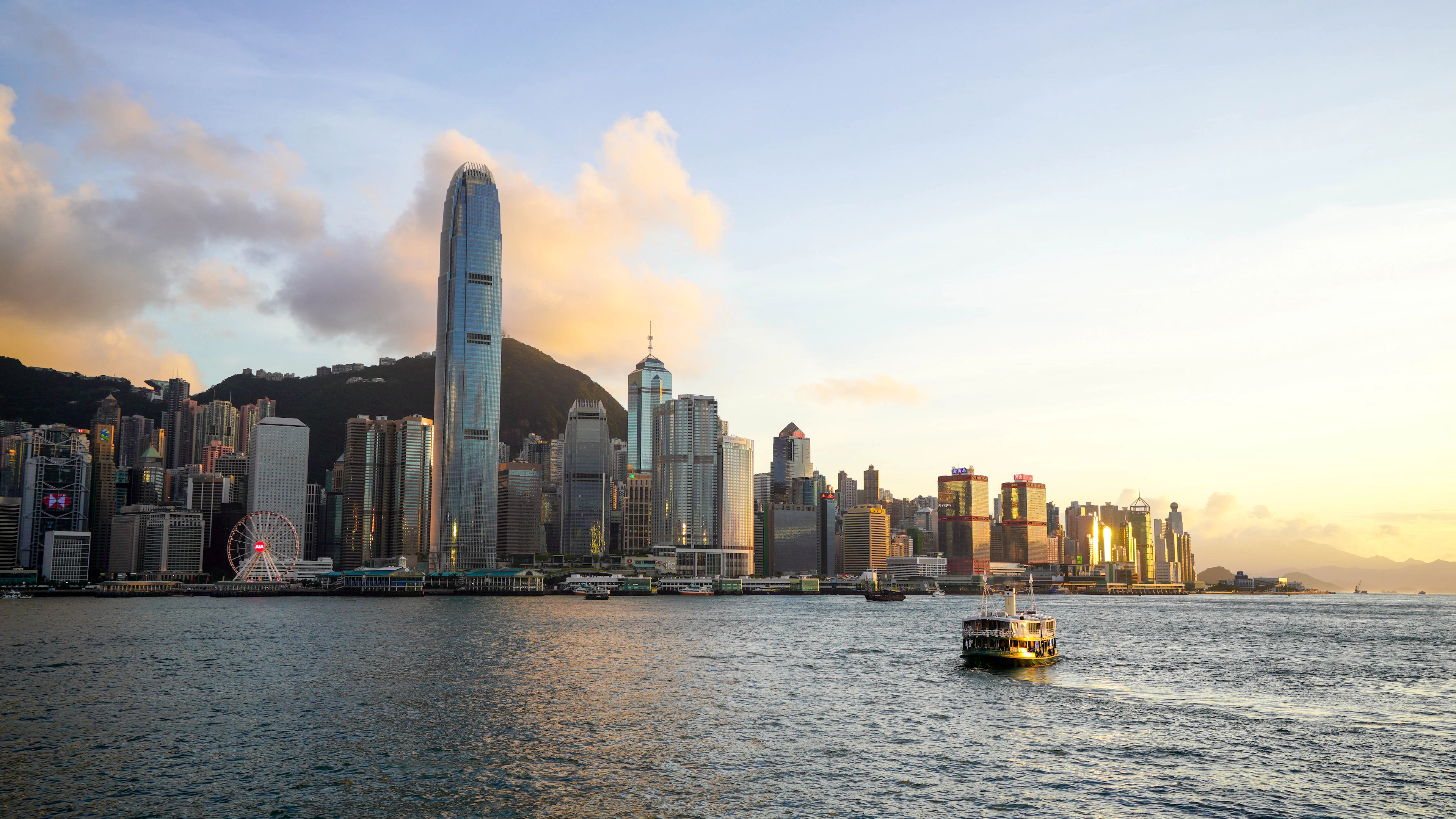 Victoria Harbour ao entardecer, com os arranha-céus da Ilha de Hong Kong e uma balsa cruzando a baía.