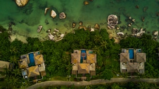 Aerial view of several thatched-roof houses surrounded by lush greenery, with blue swimming pools adjacent to each house. The houses are located near the edge of clear, shallow green water, interspersed with various-sized rocks.