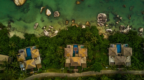Aerial view of several thatched-roof houses surrounded by lush greenery, with blue swimming pools adjacent to each house. The houses are located near the edge of clear, shallow green water, interspersed with various-sized rocks.