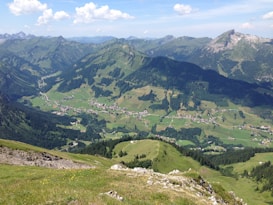 A panoramic view of a valley surrounded by towering mountains, with a picturesque village nestled in the lush green landscape. The foreground highlights rocky terrain and grassy slopes, while the background reveals mountain peaks under a bright blue sky with scattered clouds.
