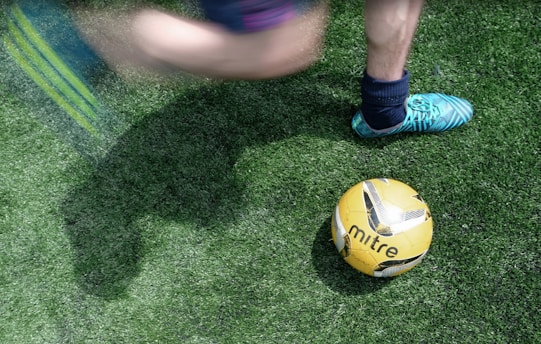 A person wearing blue and green soccer cleats is about to kick a yellow and black soccer ball on a lush green grass field. The image captures the motion of the leg swinging towards the ball, indicating imminent contact.