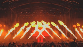 A vibrant concert scene featuring dramatic pyrotechnic flames shooting upwards from the stage. A DJ stands behind a console amidst a sea of enthusiastic fans with their hands in the air. The stage is illuminated with red and yellow lights, enhancing the fiery atmosphere.