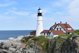 A white lighthouse is situated on a rocky shoreline with an adjacent two-story building featuring red roofs. The ocean stretches into the distance under a mostly blue sky with light clouds. Greenery is sparsely scattered among the rocks.