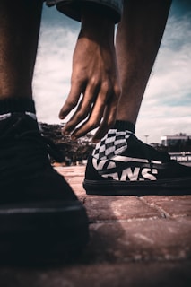 A close-up perspective of a person's hand reaching down towards a checkerboard-patterned sneaker on a brick pavement. The shoe features a prominent brand logo. The image captures a sense of movement and style, set against a slightly overcast sky.