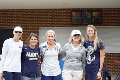 Five women wearing sports-related attire stand together, smiling in front of a brick building with a closed window. They wear various Navy shirts and hats, suggesting a team or event presence.