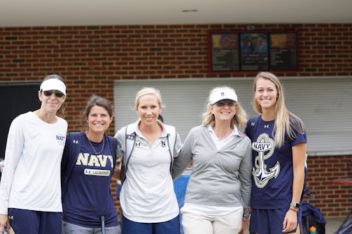 Five women wearing sports-related attire stand together, smiling in front of a brick building with a closed window. They wear various Navy shirts and hats, suggesting a team or event presence.