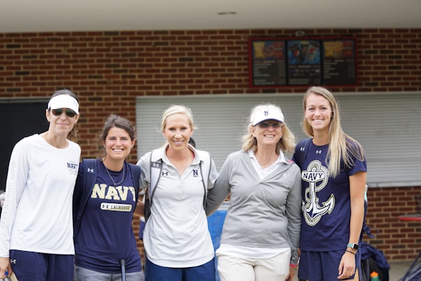 Five women wearing sports-related attire stand together, smiling in front of a brick building with a closed window. They wear various Navy shirts and hats, suggesting a team or event presence.