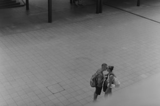 A black and white photo captures a tender moment between two people sharing a kiss in an empty, open tiled area. Both are wearing casual clothing and carrying backpacks.
