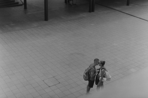 A black and white photo captures a tender moment between two people sharing a kiss in an empty, open tiled area. Both are wearing casual clothing and carrying backpacks.