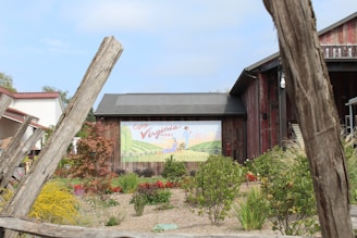 A rustic building is surrounded by a variety of plants and flowers. The central focus is a vintage-style mural promoting Virginia wines, featuring rolling hills and a figure in the distance. The wooden structure has a weathered appearance, and the surrounding garden includes vibrant green, yellow, and red foliage.