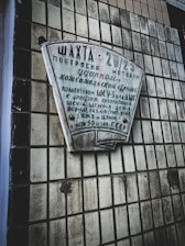 A concrete wall with a grid-like pattern of square tiles holds a plaque with Cyrillic text, commemorating the construction of a facility known as 'Шахта' in 2023 using a method described on the plaque. The text includes various Soviet references and mentions different organizations involved in construction.