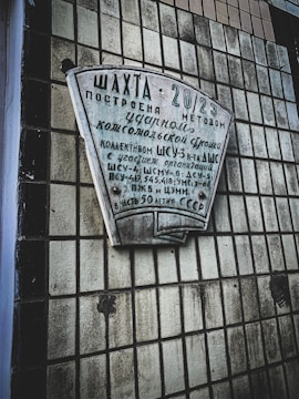 A concrete wall with a grid-like pattern of square tiles holds a plaque with Cyrillic text, commemorating the construction of a facility known as 'Шахта' in 2023 using a method described on the plaque. The text includes various Soviet references and mentions different organizations involved in construction.