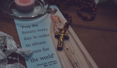 A religious pamphlet features an illustration of Our Lady of Fatima with a quote about praying the rosary for world peace. A wooden rosary with a crucifix is placed on top of the pamphlet. A pink tea light candle sits on a clear glass holder nearby, and a piece of white lace fabric is partially visible at the edge.